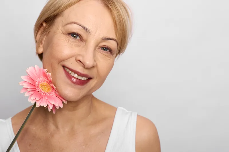 Woman portrait, smile with pink gerbera flower, soft warm senior beauty, happiness in gentle light, calm expression, studio background, wellbeing concept, fresh