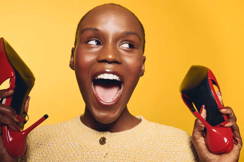 Excited black woman with dark skin holding red high heel shoes near her face on yellow background. Concept of fashion, style, shopping, and joyful emotions in studio lighting.