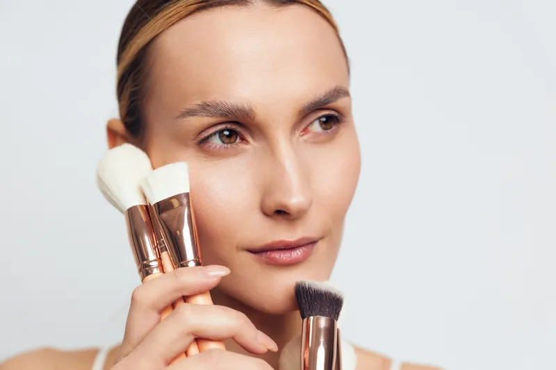 Young woman holding makeup brushes near her face, natural skin tone, soft lighting, clear background, beauty concept, minimal makeup, focused expression, clean look