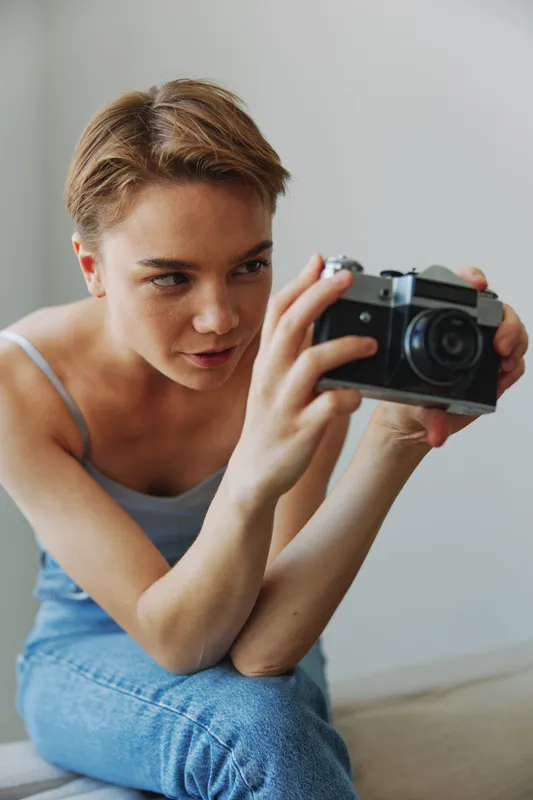 Женщина-фотограф снимает в студии на старую плёночную камеру, делая портрет на диване дома на белом фоне