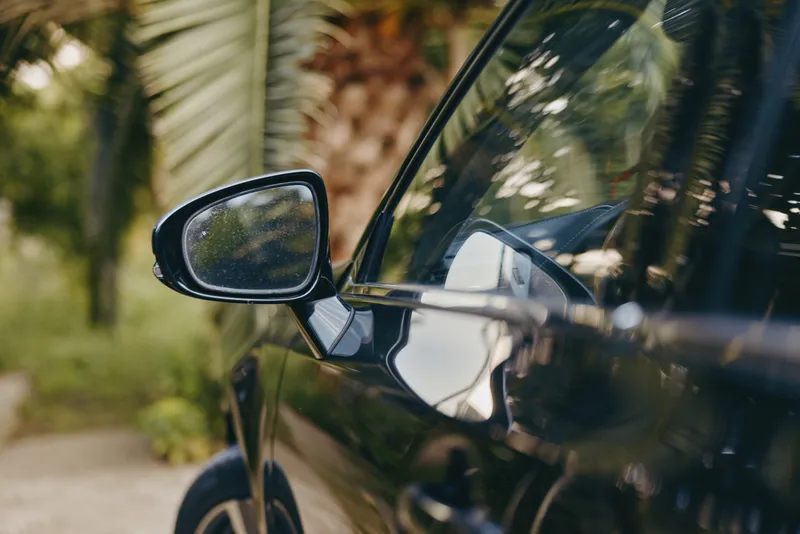 Car side mirror on a black car parked under tropical palm trees, wet droplets on glass and reflection of greenery, new luxury sedan detail shot for travel lifestyle and summer mood.
