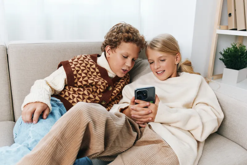 Two children sit close on a soft couch, sharing a phone screen, wearing knit sweaters and blankets, creating a warm, candid moment of curiosity and friendship.
