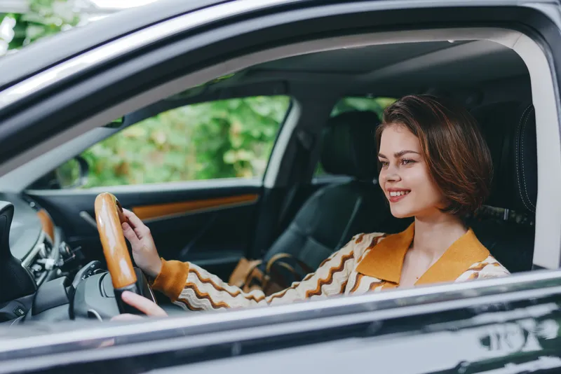 A woman is driving a car in the front seat, smiling as she grips the steering wheel, daylight streaming through the window.