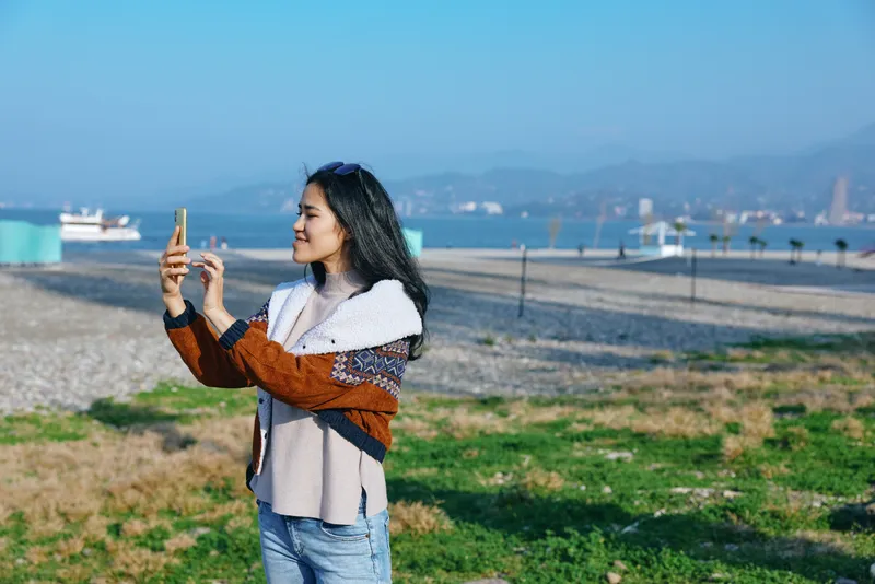 Selfie of a woman on a sunny beach holding smartphone, casual coastal fashion, sea in background, dunes and blue sky, capturing travel moment and outdoor leisure