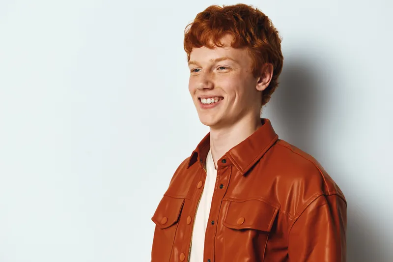 Young man with red hair and freckles smiling wearing brown leather jacket white shirt against white background studio portrait of happy confident male person.