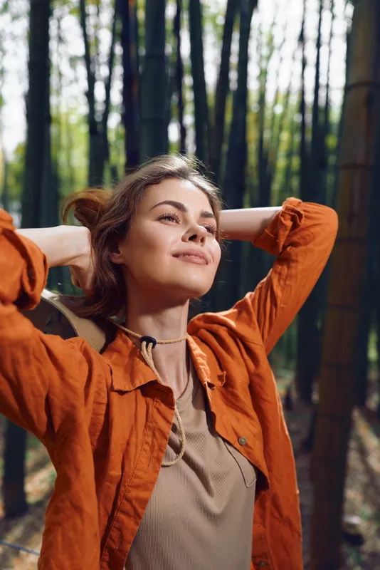 Woman portrait in a forest, relaxed and serene with sunlight filtering through tall trees. Outdoor nature scene of a smiling brunette in an orange jacket enjoying peaceful fresh air.
