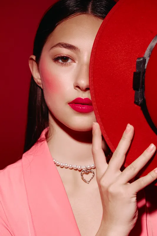 Elegant woman with red lips hiding half of her face behind a wide-brimmed red hat. She wears soft pink makeup and a rhinestone necklace. Trendy creative fashion portrait.