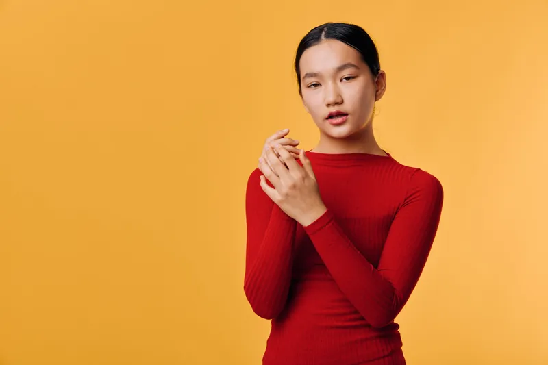 Woman in red sweater stands against orange background with hands clasped, looking thoughtfully. Portrait of young female with natural expression and casual style in studio setting.