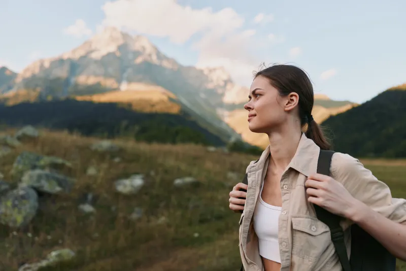 A woman on a hike pauses to admire distant mountains, wearing a light shirt and backpack. A lifestyle moment highlighting exploration, nature, and calm energy in an active daytime scene.
