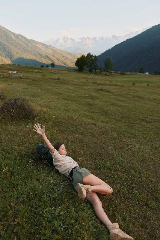 Young woman lying on green grass in a mountain valley with arms stretched, enjoying nature and fresh air during a sunny day outdoors.