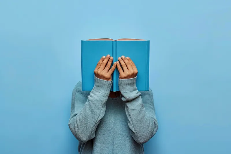 Женщина прячет лицо за синей книгой на синем фоне