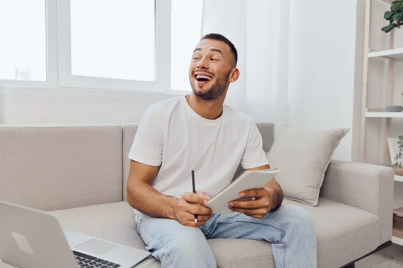 Happy man sitting on a couch, smiling joyfully while holding a notepad and pen in a bright, modern room. Perfect for concepts of creativity and inspiration.