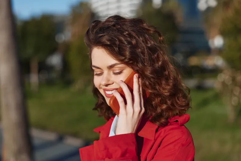 Smiling young woman with curly hair in a red jacket talks on a smartphone in a sunny park, conveying casual conversation and everyday confidence and relaxed vibe.