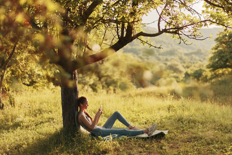Young woman enjoying a peaceful moment under a tree while using a smartphone in a sunlit meadow, evoking relaxation and connection with nature