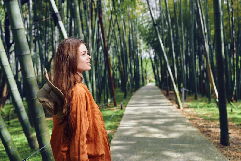 Woman traveler with hat and orange coat standing on a bamboo path in a peaceful forest, looking along the trail for exploration and quiet nature walk, scenic outdoor portrait.