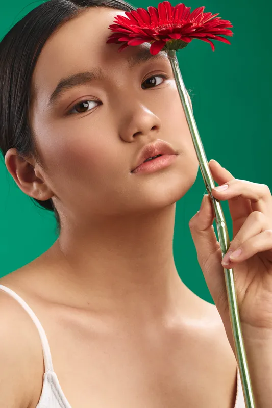 asian woman gazes softly at the camera, holding a red flower against a green backdrop, creating a serene portrait with gentle lighting and natural composition.