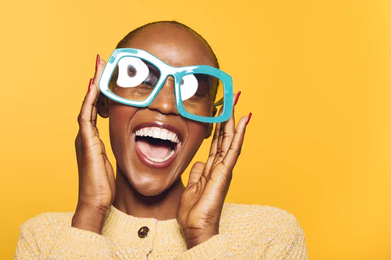 Happy African American woman with dark skin wearing oversized blue glasses smiles widely on yellow background. Concept of joy, fashion, and positive lifestyle with vibrant colors and energy.