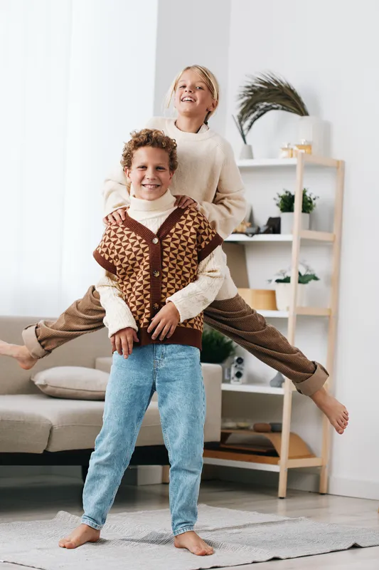 Two children play indoors, with a cheerful girl perched on a boy's back, smiling and balancing gracefully. Cozy sweaters and denim create a warm, joyful scene.