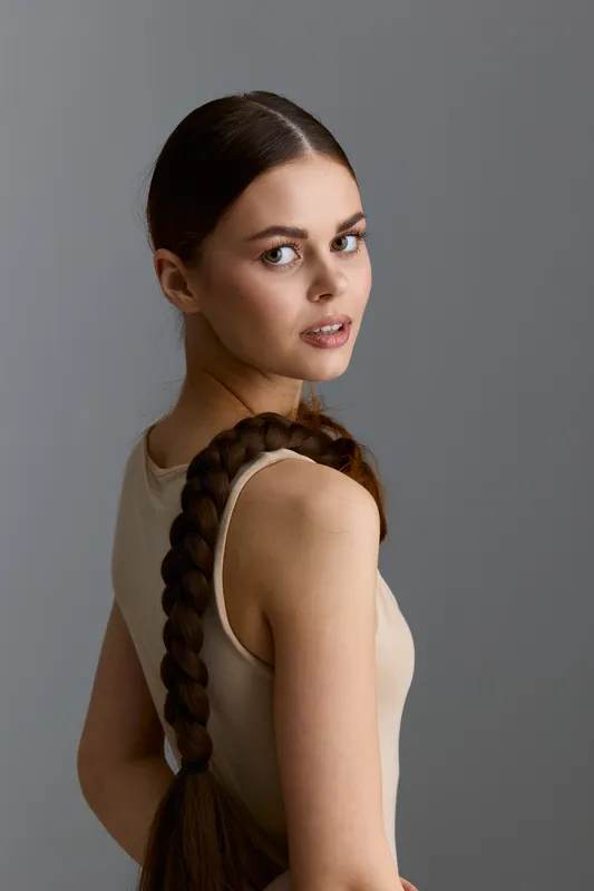 young woman with long braided hair wearing a beige sleeveless top looking over shoulder against neutral gray background in soft natural lighting