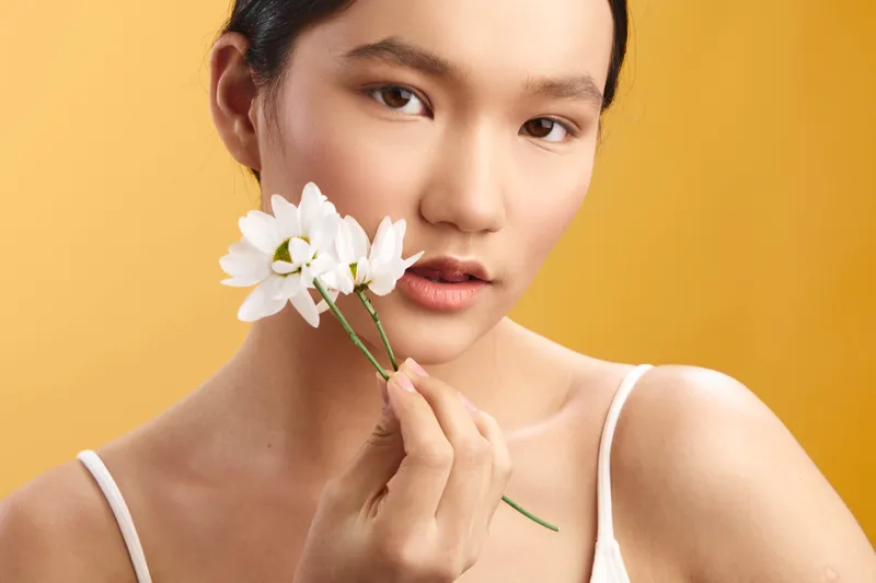 Asian woman holds a white flower near her lips in a warm studio setup, soft glow emphasizes smooth skin and a serene, elegant mood.