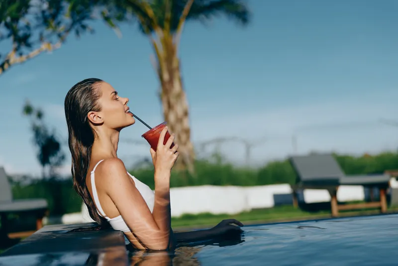 A woman relaxes in a pool, sipping a red drink through a straw in bright outdoor light. A lifestyle moment of leisure, hydration, and summer refreshment.