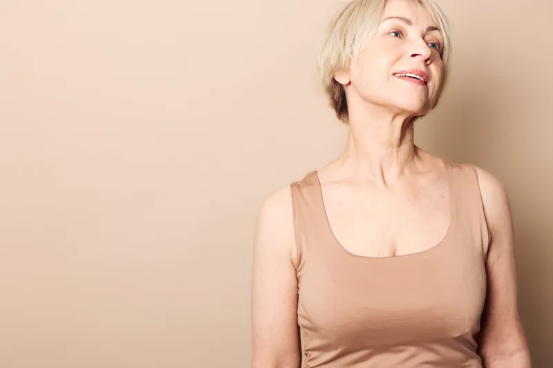 Portrait of a mature woman with short blonde hair wearing a beige tank top, looking away with a soft smile. Neutral background enhances the calm and natural mood of aging beauty.
