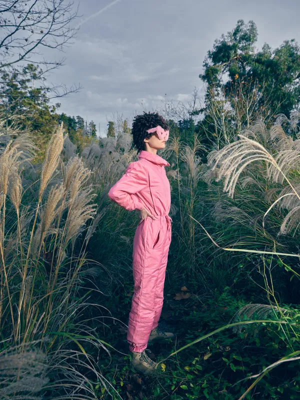 Pink jumpsuit worn by a woman in an outdoor landscape, a fashion portrait among tall grasses, wearing a mask, showcasing bold color and natural textures.