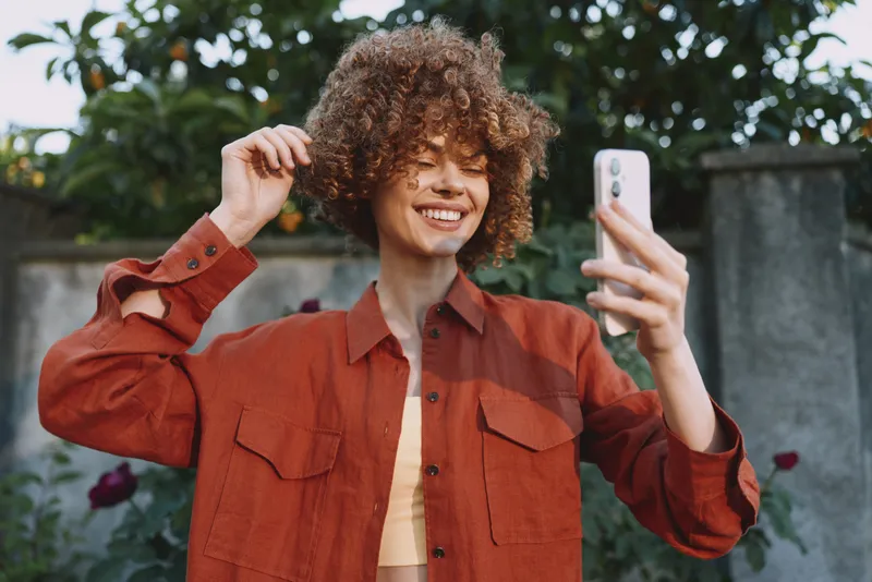 Woman lifestyle selfie outdoors smiling in a rust jacket, casual fashion portrait with natural light and urban vibe, candid confident pose, vibrant colors, cheerful mood and energy