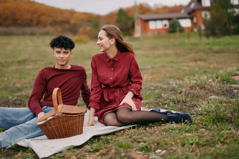 Young couple sits on a blanket in an autumn meadow sharing a quiet picnic with a wicker basket; candid, natural poses and genuine smiles emphasize authenticity and realistic everyday connection.