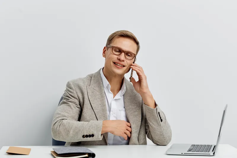 Professional smiling man talking on the phone while working at his office desk, wearing glasses and a gray checkered blazer, with a laptop and notebooks on white background, business communication con