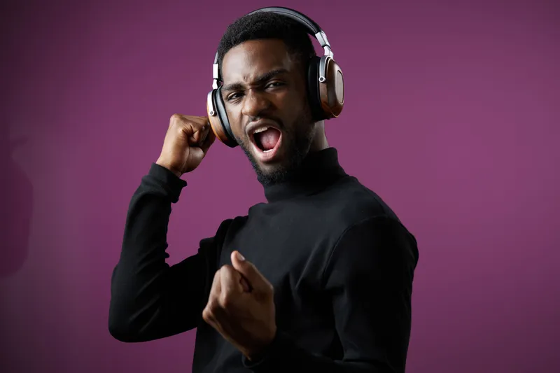 Young man celebrating with headphones, wearing a black turtleneck in front of a vibrant purple background, expressing joy and energy in a trendy lifestyle shot