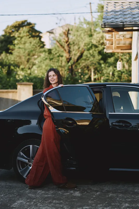 Cheerful woman in a red dress outdoors beside a sleek car, sunlit moment, relaxed pose, casual lifestyle vibe, vibrant colors with green foliage in the background