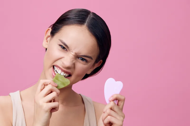 Young woman demonstrating skincare routines with gua sha tool against a pink background, showcasing emotions of determination and self care