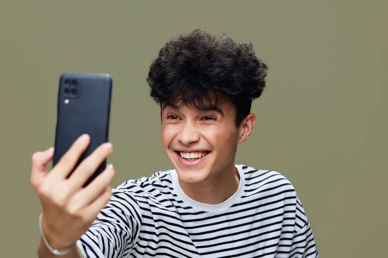 smiling man taking selfie with smartphone, casual striped shirt, curly hair, joyful expression, indoor neutral background, modern lifestyle portrait.