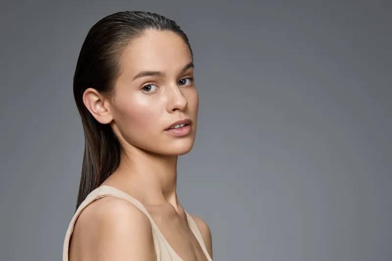 Elegant young woman with smooth skin and wet hair, showcasing a natural beauty look against a gray background, emphasizing simplicity and confidence