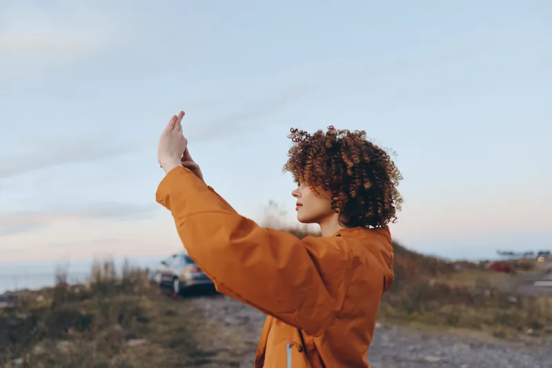 Woman lifestyle portrait outdoors at sunset on beach dunes, curly haired woman raises one hand in a friendly wave, warm jacket and soft light, calm mood windy sky and distant shoreline