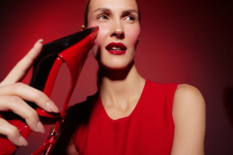 Woman in red dress holds red high heel shoe near face on red background. Fashion concept with bold makeup and stylish accessories in studio lighting.
