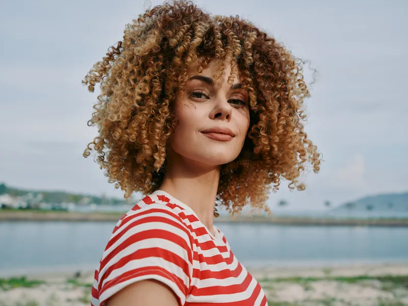Portrait of a confident woman with voluminous curly hair wearing a red and white striped shirt, standing by a calm coastal backdrop under a soft sky.