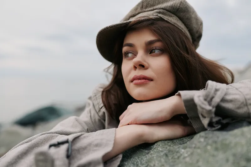 Thoughtful young woman wearing a cap leans on a rock outdoors. She looks to the side with a calm expression under overcast sky. Concept for contemplation, nature, and casual fashion.