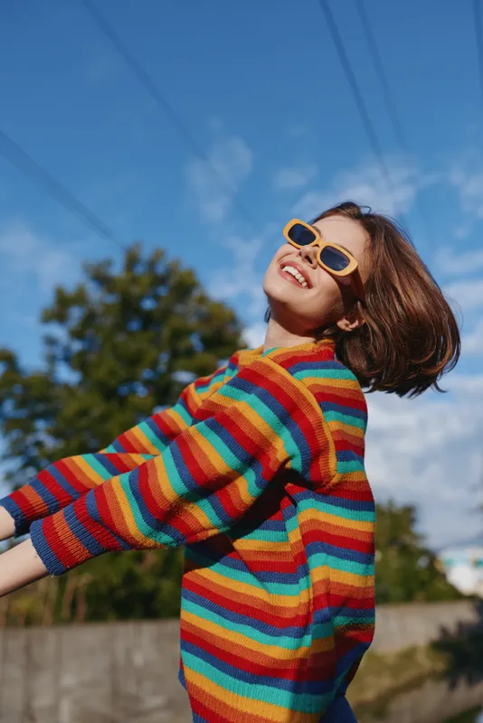 Young woman in a rainbow sweater and sunglasses, smiling outdoors on a sunny day; short brown hair, leaning back and laughing in a colorful casual outfit for lifestyle fashion.