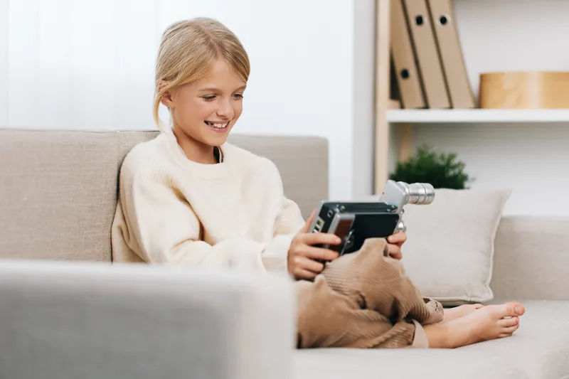 A young girl sits barefoot on a cozy sofa, smiling as she holds a small handheld camera, capturing a playful moment in a bright, tidy living room.
