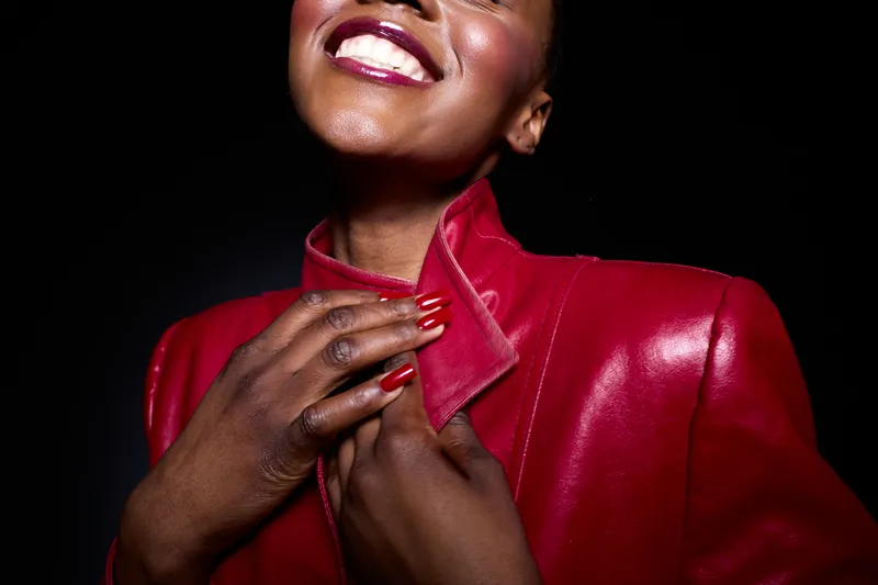 Smiling black woman with dark skin wears red leather jacket and matching nail polish on black background. Concept of fashion, beauty, confidence, and elegance in studio portrait.