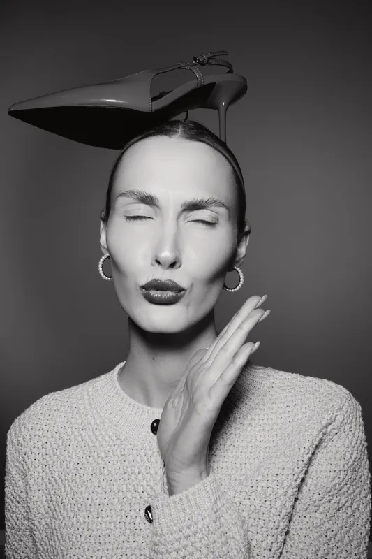 Portrait of light skinned Caucasian woman with eyes closed and lips pursed, wearing hoop earrings and textured sweater, balancing high heel shoe on head in studio.