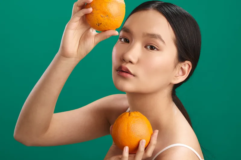 Asian woman holds bright oranges against a vivid green background, close up portrait with soft lighting, calm expression, contemporary citrus themed still life portrait.