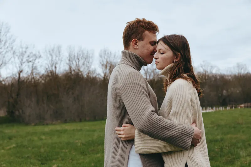 Romantic couple embraces in a park and shares a kiss, wearing oversized sweaters, calm natural light, soft autumn tones, intimate lifestyle moment, candid outdoor affection.