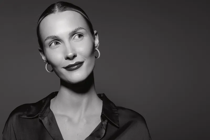 Portrait of a young white woman with pale skin and slicked back hair wearing hoop earrings and a satin shirt looking sideways on dark background.