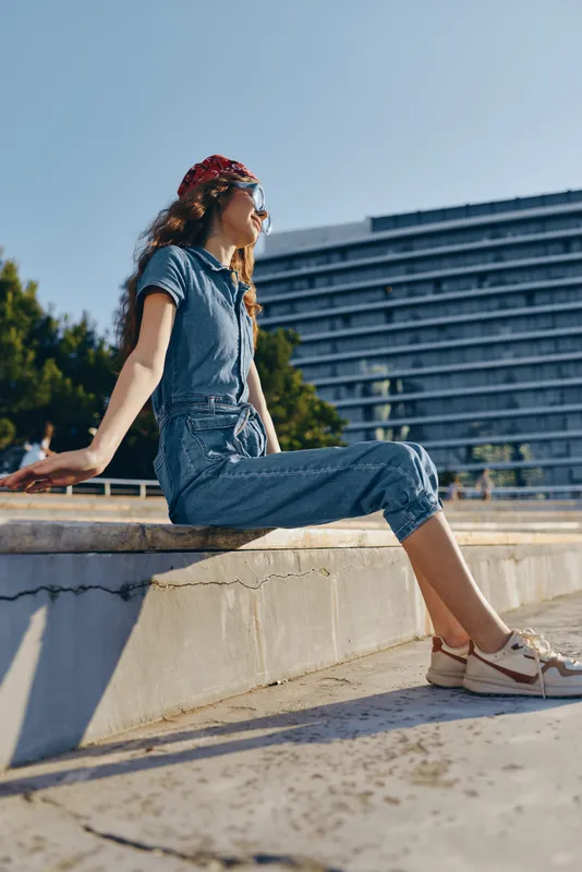 Lifestyle woman in modern boho-western denim street style relaxing outdoors under clear blue sky with film color effect and casual sneakers.