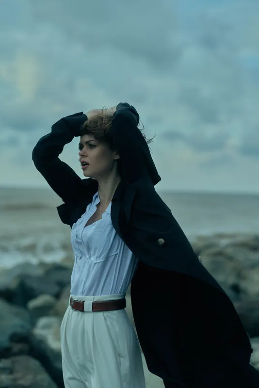 Woman on a windy beach wearing a dark coat draped over a light shirt and white trousers, arms raised, dramatic movement against a rugged seascape, outdoor fashion mood