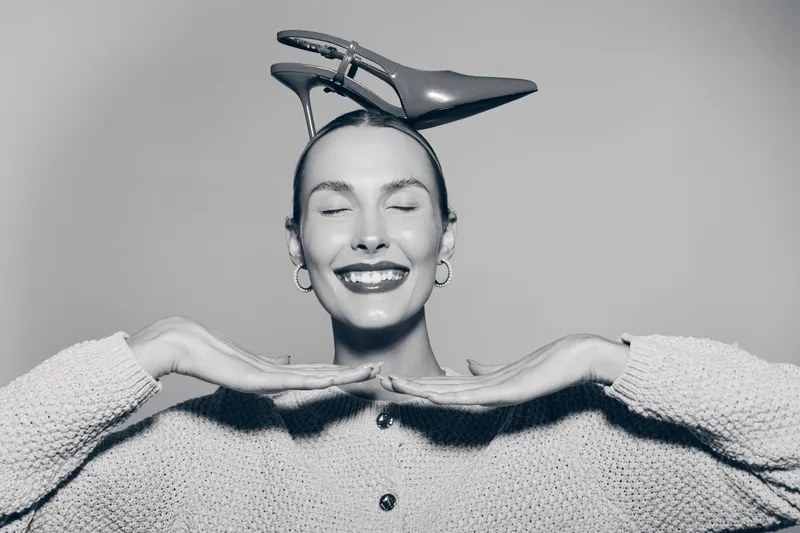 Portrait of a smiling young white woman with closed eyes wearing a knitted sweater and holding a high heel shoe on her head. Concept of happiness, fashion, and playful mood.