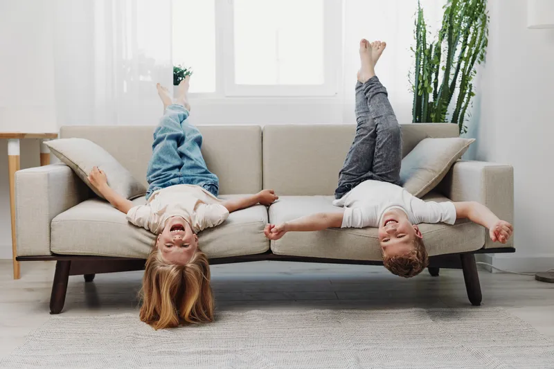 Two happy children lying upside down on a sofa in a bright living room, laughing and holding hands. Concept of friendship and joy.